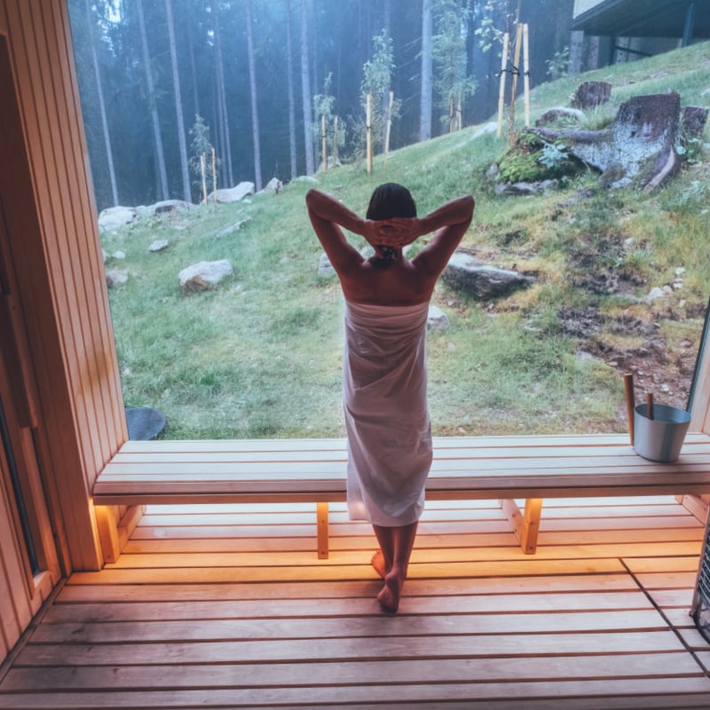 Relaxing Woman wrapped white towel in Hot Finnish sauna with a huge wide window enjoying the green forest