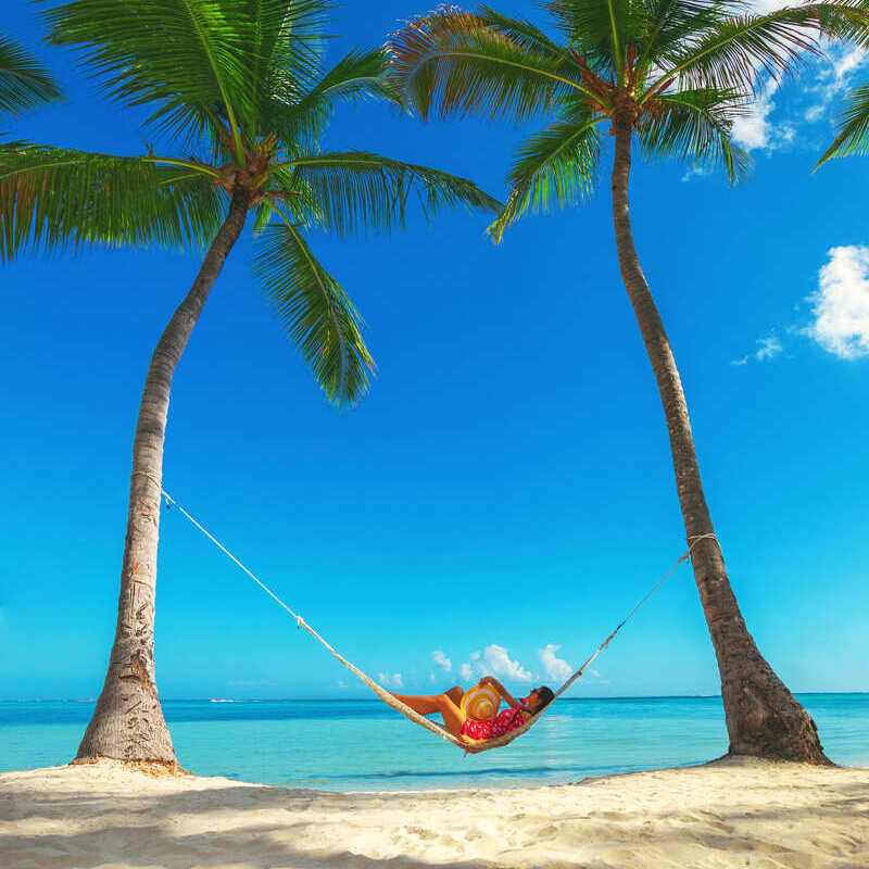 A Young Woman Resting On A Hammock Suspended Between Two Tall Palm Trees In Punta Cana, The Dominican Republic, Latin America