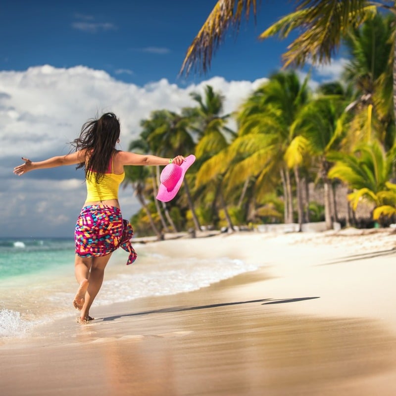 A Young Woman Running On A Sandy Beach Bounded By Tall Palms In Punta Cana, The Dominican Republic, Latin America