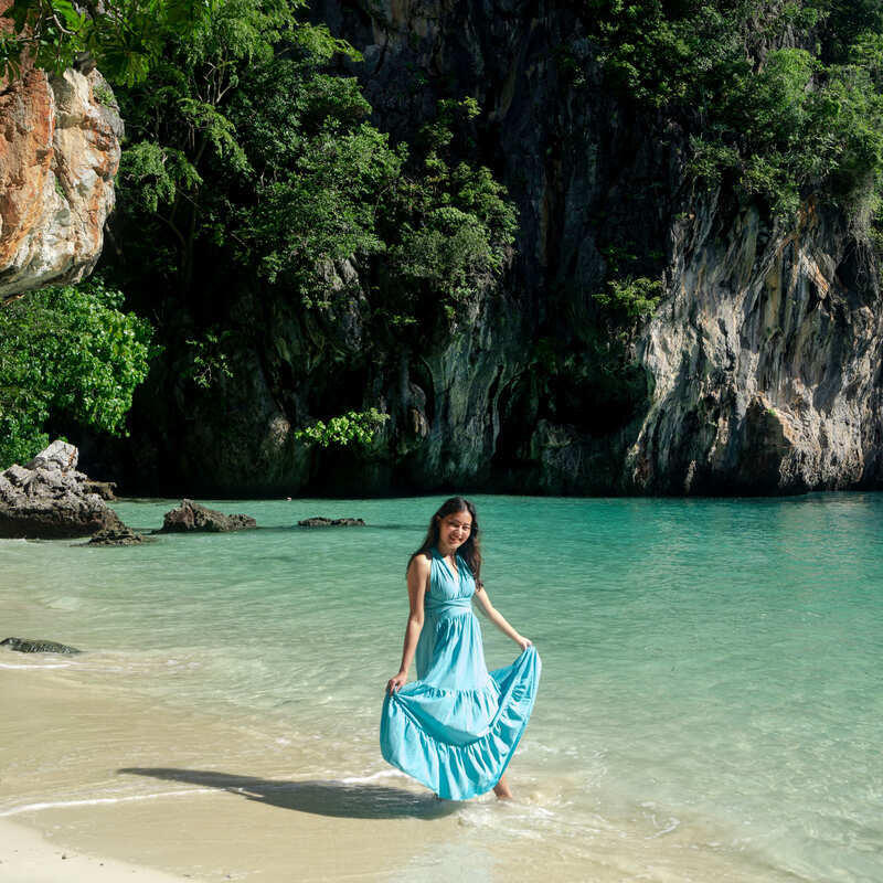 Young Female Tourist Wearing A Blue Dress And Smiling As She Walks Lao Lading Beach In Thailand, Southeast Asia