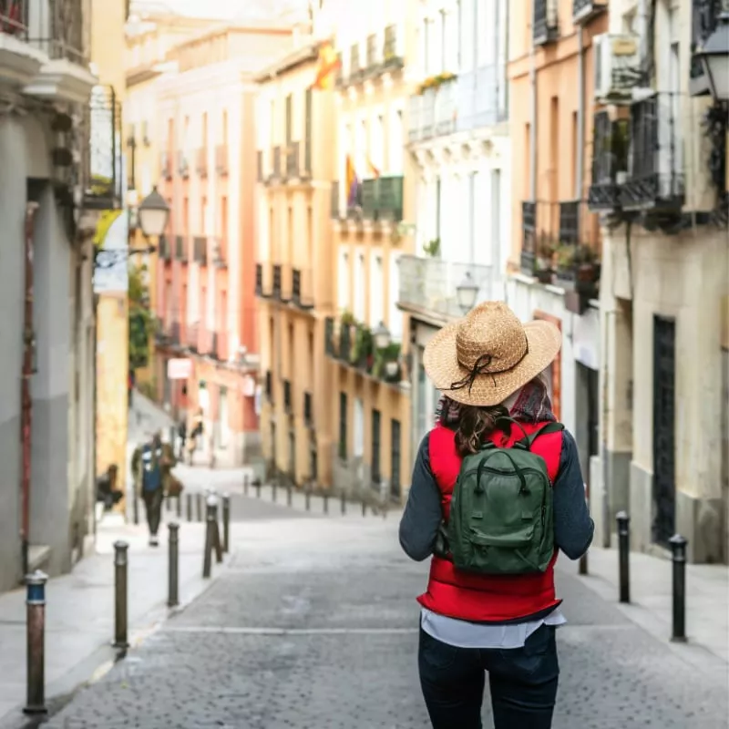 A female tourist with her back turned, wearing a hat and backpack, walks down a street in the historic center of Madrid. Hiking tourism in Spain. Concept of Spanish tourism and culture.