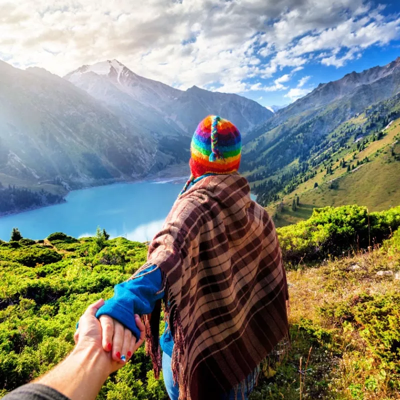 Tourist woman in rainbow hat and brown poncho holding man by hand and going to the lake in the mountains in peru