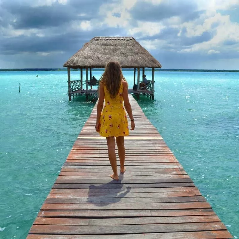 Young Woman In A Yellow Dress Walking A Pier Stretching Out Onto Lake Bacalar, Mexican Caribbean, Mexico