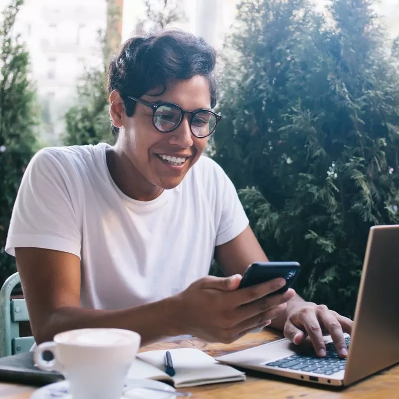 Smiling Remote Worker, Digital Nomad Checking His Phone As He Works From His Computer In A Cafe Setting, Unspecified Location