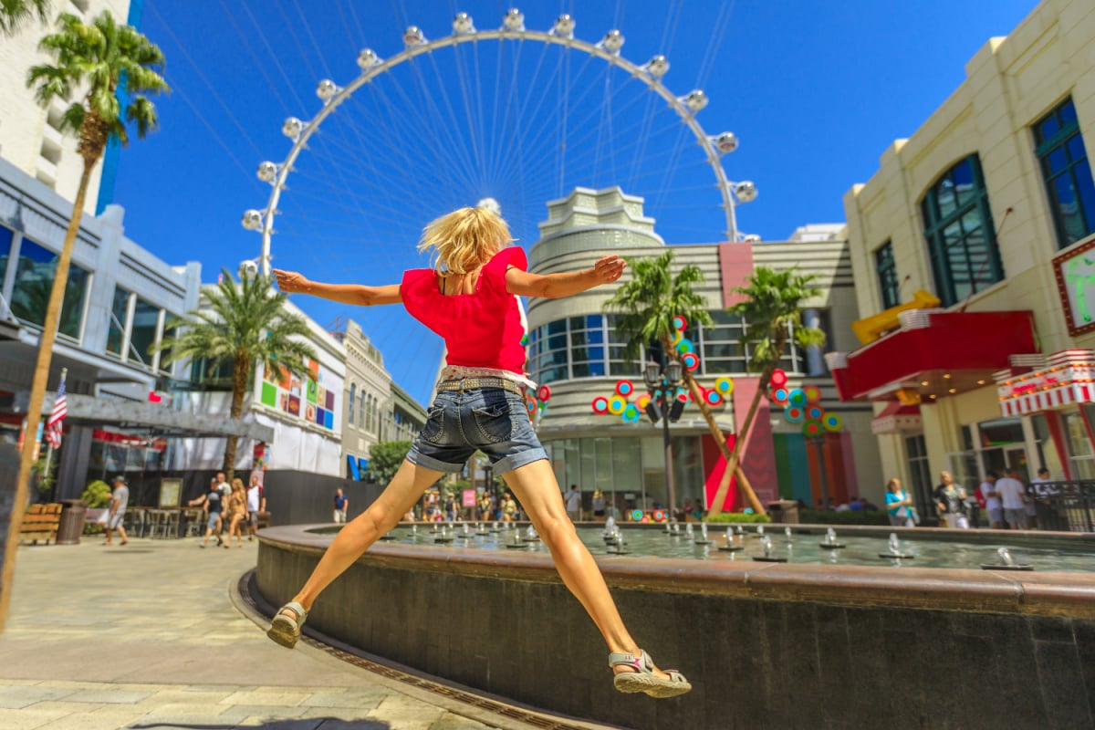 Blonde woman jumping at center of Las Vegas in a sunny day, blue sky. Ferris Wheel, the world's largest observation wheel on blurred background. Happy tourist in Las Vegas cityscape, Nevada, USA