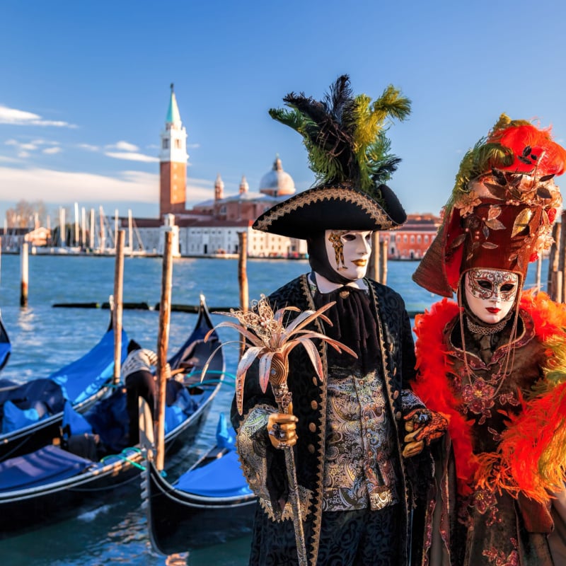 Two people in festive costumes stand in front of a canal with boats in Venice during Carnival