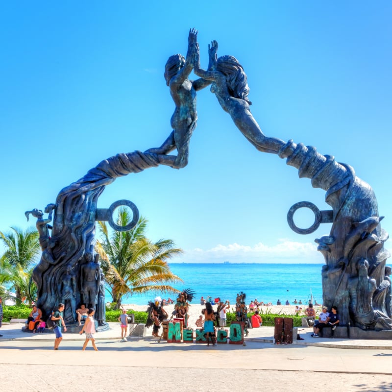 Visitors mingling on Fundadores Park beach at Playa del Carmen on the Caribbean coast of Riviera Maya with performers under the Portal Maya sculpture.