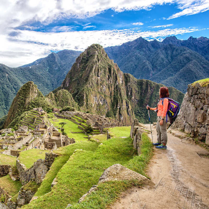 A Female Tourist Leaning On A Trekking Pole As She Admires The View Of Machu Picchu Surrounded By Green Peaks In Peru, South America