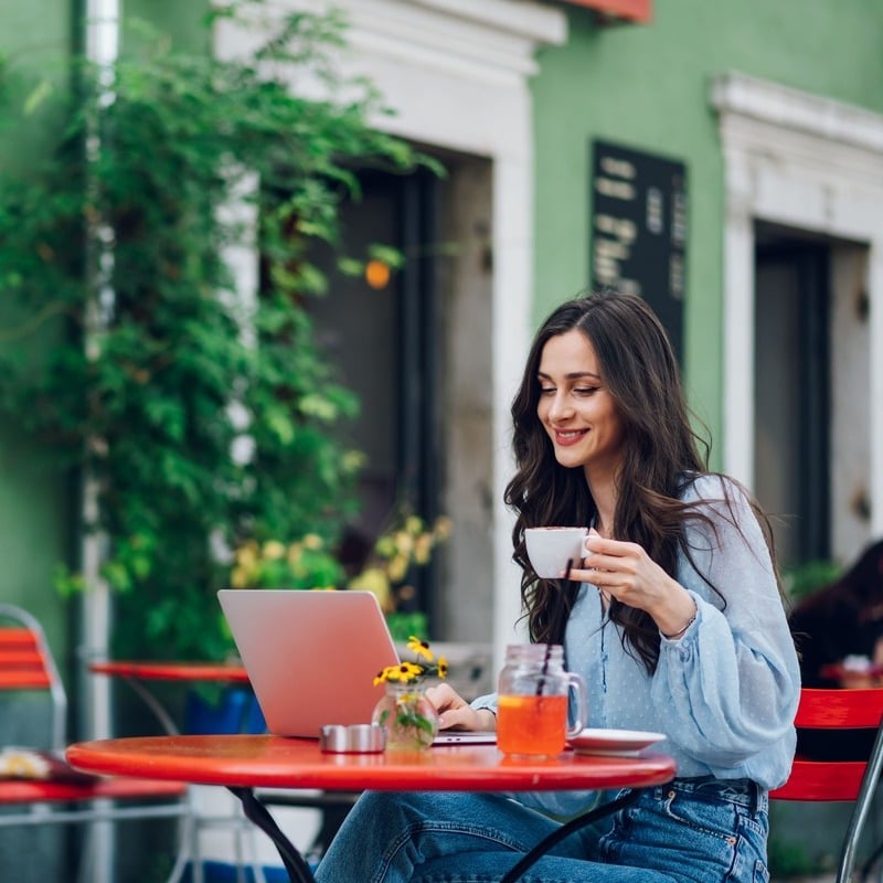 Smiling Female Digital Nomad, Remote Worker Working From Her Computer In An Alfresco Cafe As She Ships On Some Tea Or Coffee, Unspecified Location