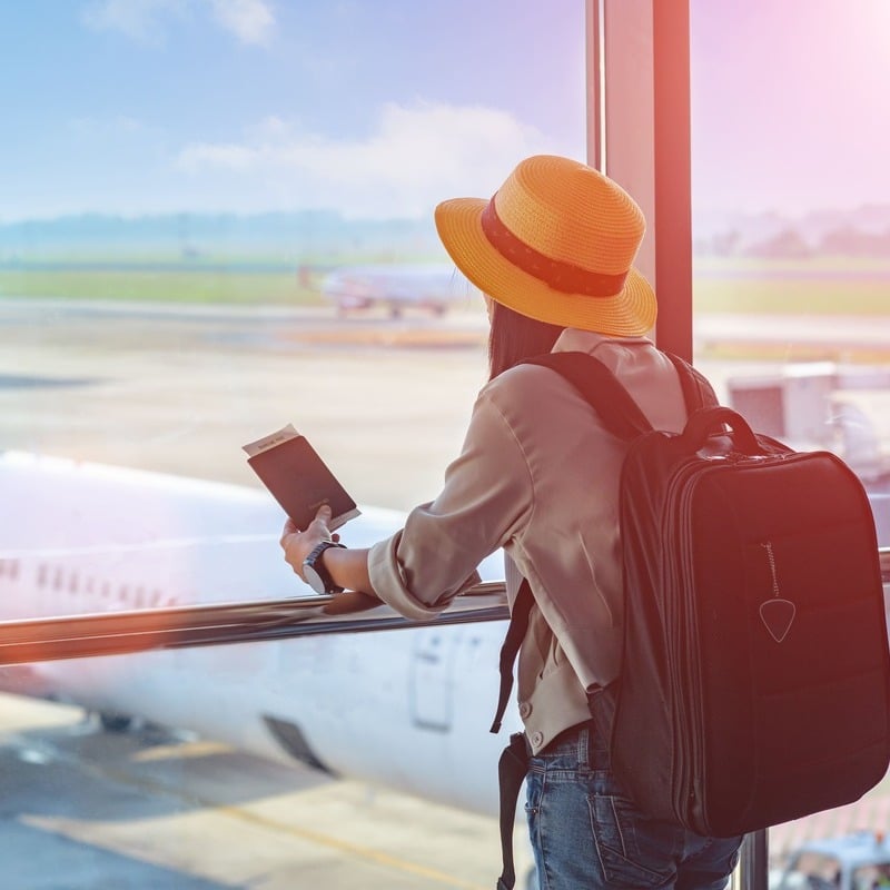 Female Traveler Observing Planes Approaching The Tarmac As She Holds Her Passport In An International Airport