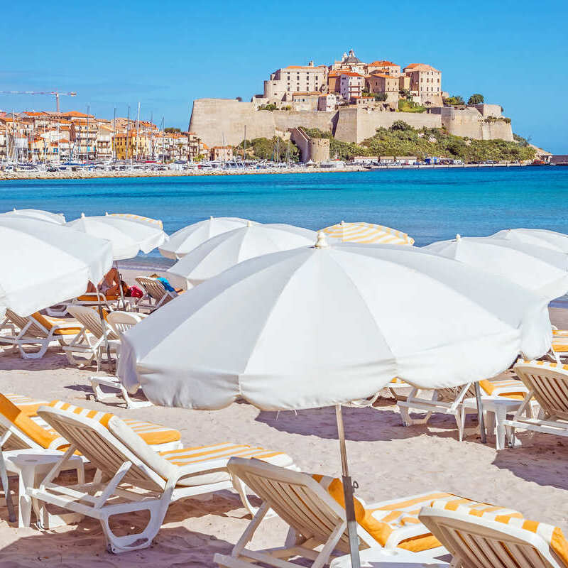 A Beach Filled With Loungers With A View Of A Historic Fortress In Corsica, France, Mediterranean Sea