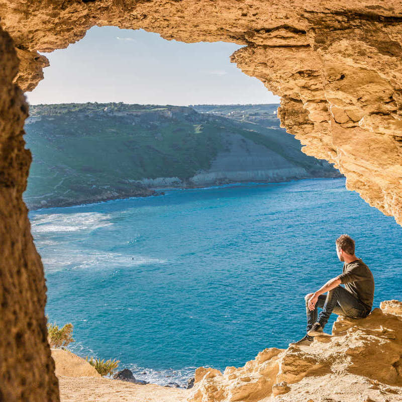 A Young Male Tourist Observing The Mediterranean Sea From A Cave Window In Gozo, Malta, Southern Europe