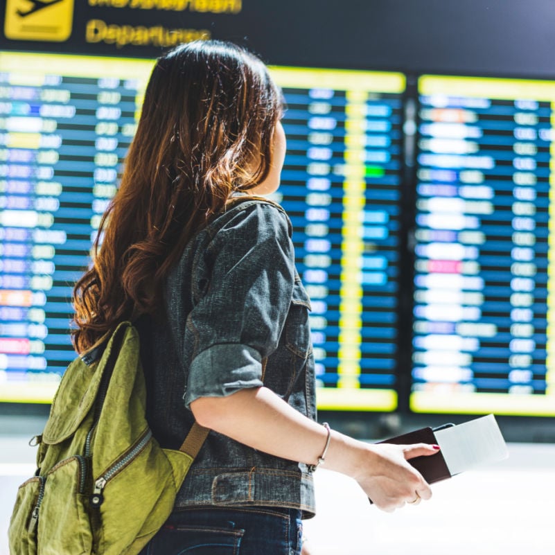 young female traveler looks at departures board at an airport while holding passport and boarding pass