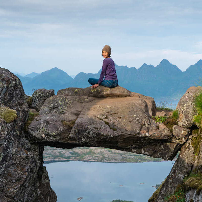 A Young Female Tourist Posing For A Picture In Djevelporten, Lofoten, A Suspended Rock Between Two Cliffs, Scandinavia, Northern Europe