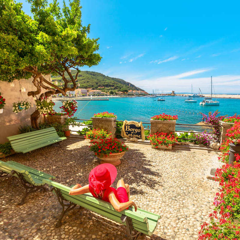 Young Woman Sitting On A Beach As She Admires A View Of The Mediterranean In The Island Of Elba, Italy, Southern Europe