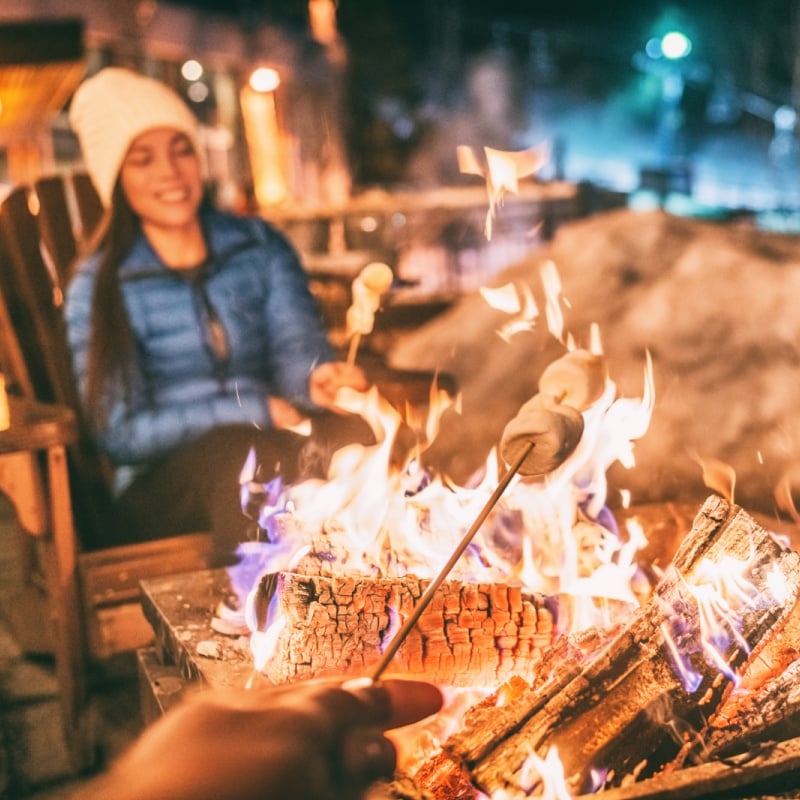 Winter holiday ski resort woman roasting marshmallows in BBQ firepit afterski fun leisure activity with friends. Couple grilling marshmallow stick in fire.