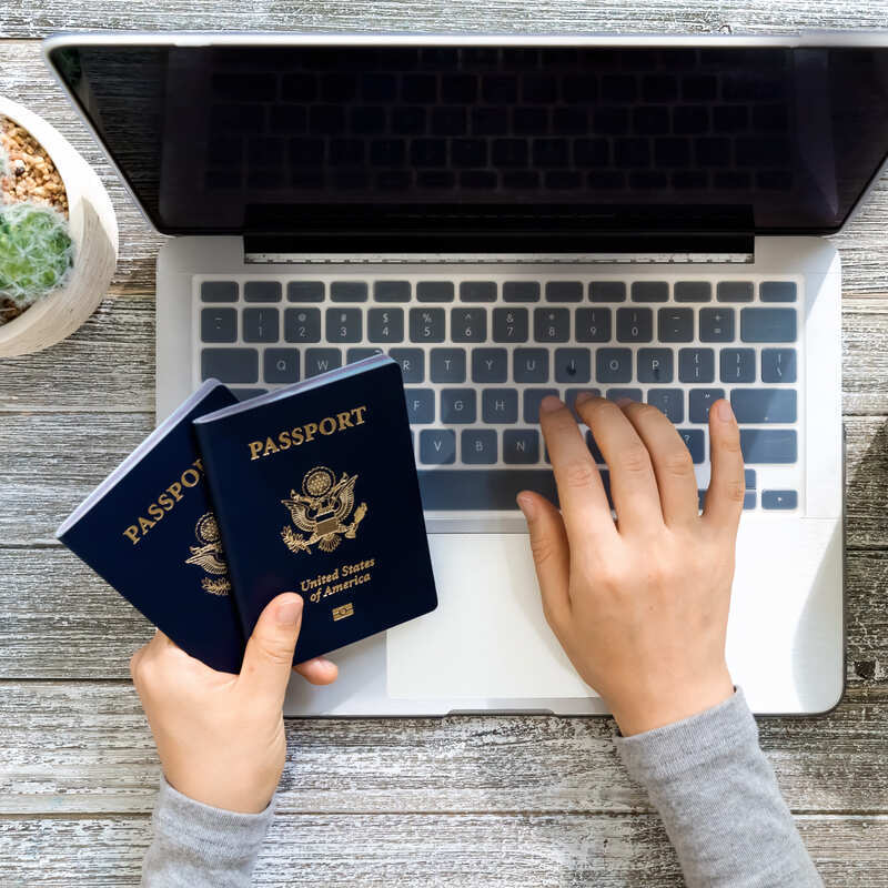 American Traveler Holding Up Two US American Passports As He Uses His Computer, Booking A Plane Or Checking Into A Flight